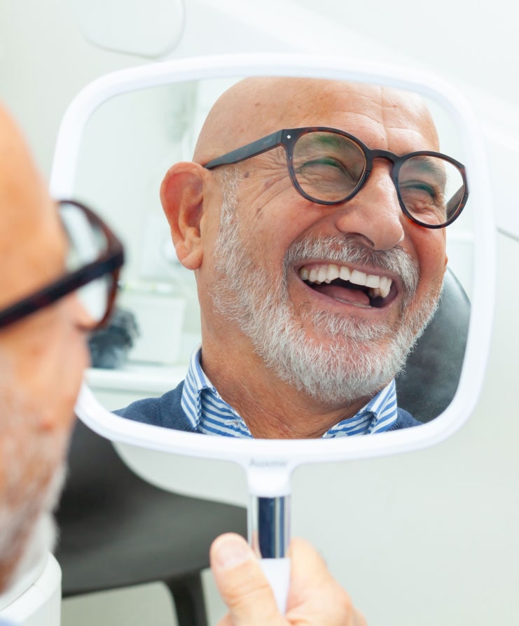 Older man smiling while looking in mirror at dental office after successful treatment